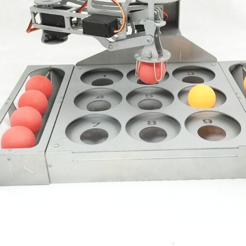 Metal teaching robot gripper holding a red ball over a numbered tray
